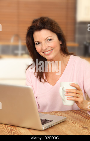 Attraente donna matura utilizzando un computer portatile, seduto al tavolo della cucina tenendo una tazza bianca, sorridente alla telecamera. Foto Stock