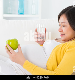 Nutrizione Materna. Asian donna incinta stile di vita sano, azienda frutta e acqua bicchiere seduti sul divano di casa. Foto Stock