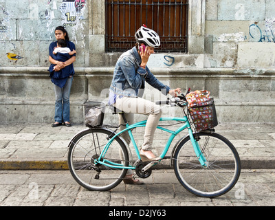 Moderno e giovane donna messicana sulla bicicletta con cellulare casco & indigeni adolescente indiano con il bambino sul marciapiede Oaxaca Foto Stock