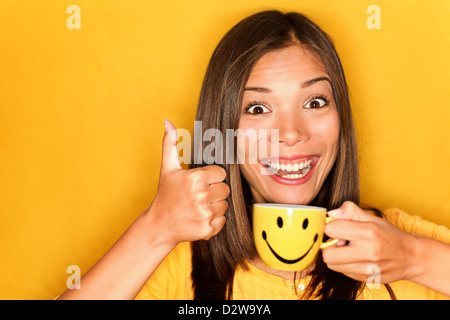 Ritratto di giovane e bella multirazziale caucasico Cinese / donna asiatica di bere il caffè dando pollice in alto su sfondo giallo Foto Stock