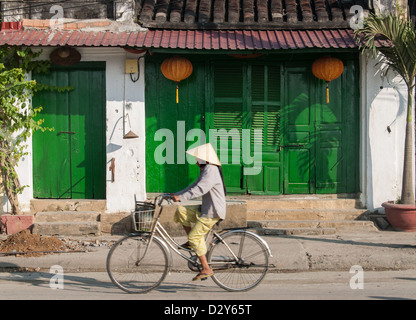 Una donna non identificato cavalca la sua bicicletta davanti ad un negozio chiuso con porte verdi su gennaio 10, 2008 in Hoi An, Vietnam. Foto Stock