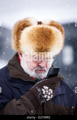 Senior uomo nel cappello di pelliccia in piedi nella neve e azienda smart phone Foto Stock