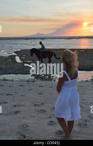 Una donna con abito bianco guarda un cavaliere e il tramonto su Bali in Gili Trawangan; Lombok, Indonesia. Foto Stock