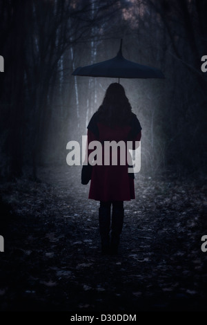 Una donna con un cappotto rosso e un ombrello è a piedi attraverso una foresta scura Foto Stock