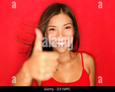 Ritratto di Allegro e gioioso multirazziale Asian / donna caucasica mostrando pollice in alto segno positivo su sfondo rosso Foto Stock