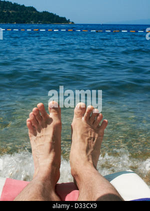 Gambe e piedi dell'uomo su una sedia da spiaggia con vista mare. Villaggio di Torba, penisola di Bodrum, Turchia. Foto Stock