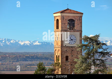 Vista sui mattoni vecchi torre campanaria con un grande orologio e montagne con cime innevate sullo sfondo sotto il cielo blu in Piemonte, Italia. Foto Stock