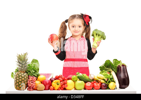 Una ragazza sorridente con grembiule tenendo un broccoli e apple rosso dietro un tavolo pieno di frutta e verdura isolato su bianco Foto Stock