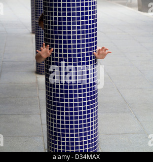 Ragazzo giovane si nasconde dietro un blu mosaico piastrellato pole che mostra solo le sue mani creando un illusione Foto Stock