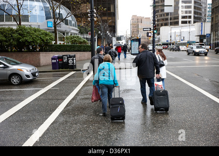 I turisti a piedi attraverso crosswalk su bagnato Rainy day tirando Borsoni con ruote Vancouver BC Canada Foto Stock