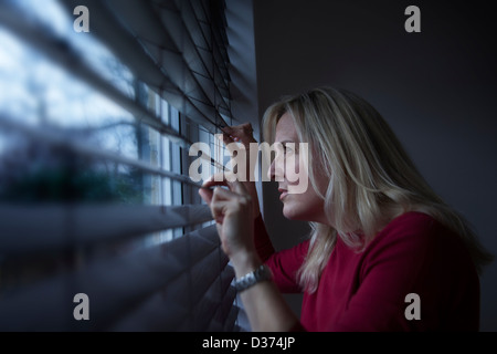 Profilo di una donna con capelli lunghi biondi, il peering attraverso una finestra cieco dall'interno guardando fuori, luce sta colpendo il suo volto. Foto Stock