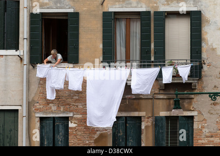 Donna con mollette nella sua bocca appendere fuori fogli bianchi e altra biancheria da letto a secco da un balcone Foto Stock