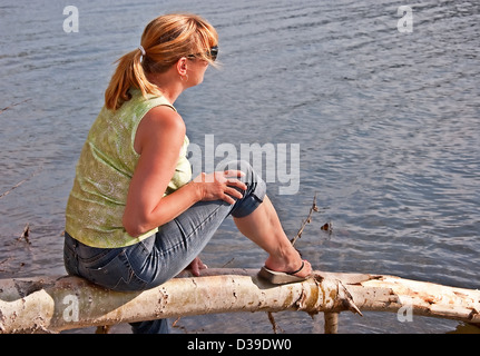 Caucasian donna con capelli biondi in una coda di cavallo, è seduta su un registro e un momento di relax a un lago. Lei sta godendo di un senza stress. Foto Stock