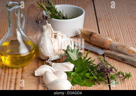 Aglio, prezzemolo, salvia, rosmarino e timo con olio extra vergine di oliva in una brocca, una spazzola e un coltello. su un antico tagliere. Foto Stock