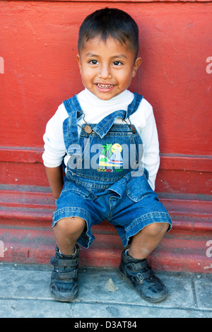 Maliziosa bright eyed sorridente piccolo messicano ragazzo si siede fuori contro la profonda parete rossa su una strada in Oaxaca de Juarez Mexico Foto Stock