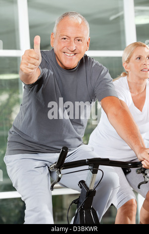 Felice l'uomo senior tenendo i pollici fino a spinning bike nel centro fitness Foto Stock