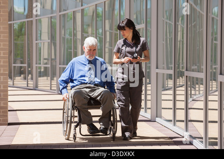 Uomo con la distrofia muscolare e il diabete nella sua sedia a rotelle a piedi con un infermiere utilizzando smart phone Foto Stock