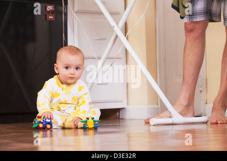 Baby boy giocando con i giocattoli sul pavimento mentre il padre è la stiratura Foto Stock