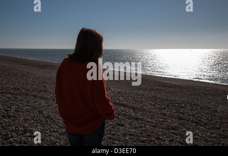 Torna alla telecamera silhouette di una donna che indossa un chunky uomini il ponticello che guarda al mare a guardare il sole barlume sull'acqua. Foto Stock