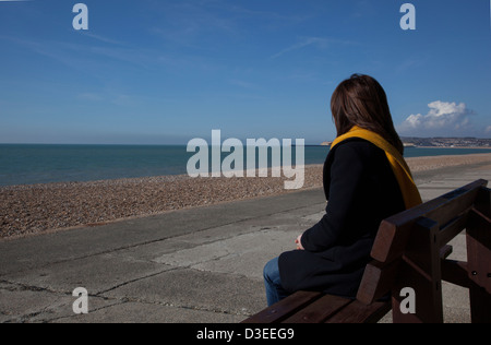 Torna alla telecamera colpo di una donna Cappotto e sciarpa seduto da solo guardando al mare, a significare la perdita di una persona cara. Foto Stock