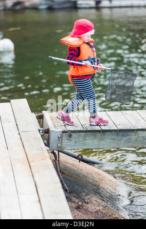 Un giovane bambino che indossa il giubbotto di salvataggio camminando sul pontile sul mare in possesso di una rete da pesca in mano Foto Stock