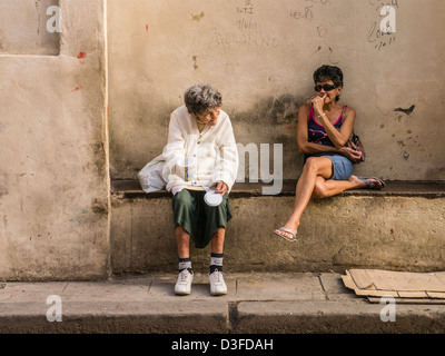 Due donne, una anziana sedersi sulla parete di cemento banco di lavoro come quello più vecchio mendica per denaro a l'Avana, Cuba. Foto Stock