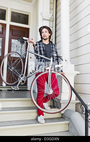 Hipsters e beatnik - un giovane uomo tanga in jeans stretti, plaid shirt e hat circa per lasciare la casa nel suo fixed-gear bike. Foto Stock