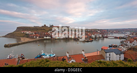 La cittadina di Whitby nel North Yorkshire guardando sopra i tetti della città attraverso il porto verso l'Abbazia rovine al tramonto Foto Stock