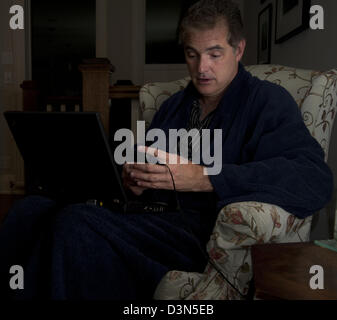Uomo seduto al buio nel suo soggiorno di notte con un computer portatile davanti a lui e mobili in mano i messaggi di testo Foto Stock