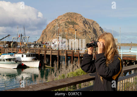 Una giovane ragazza a guardare la fauna selvatica con un binocolo in il californiano città costiera di Morro Bay con Morro Rock in background Foto Stock