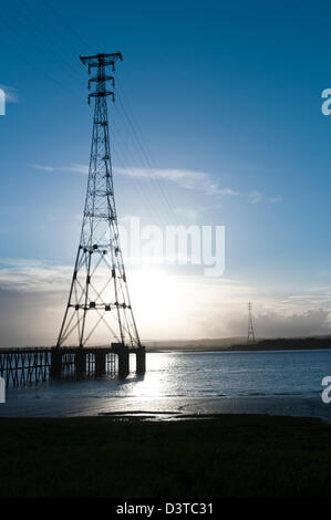 Tall tralicci elettrici a Aust portando ad alta energia linee elettriche attraverso il fiume Severn con un basso sun che si riflette sull'acqua. Foto Stock