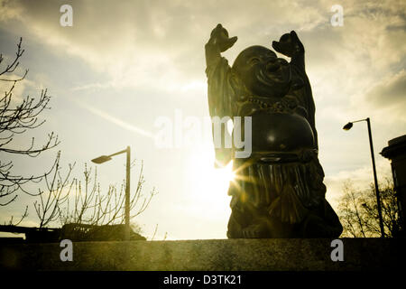 Il Buddha con le braccia in aria seduto su una mensola con luce del sole dorato proveniente da dietro e i lampioni in background Foto Stock