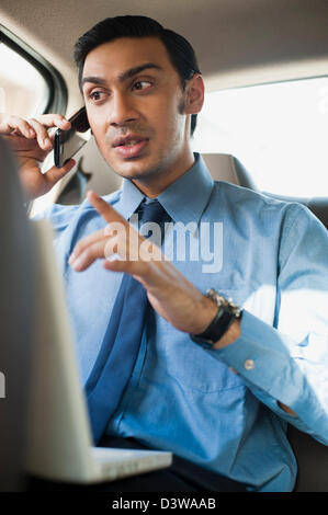 Imprenditore Bengali usando un computer portatile e a parlare su un telefono cellulare in un'automobile Foto Stock