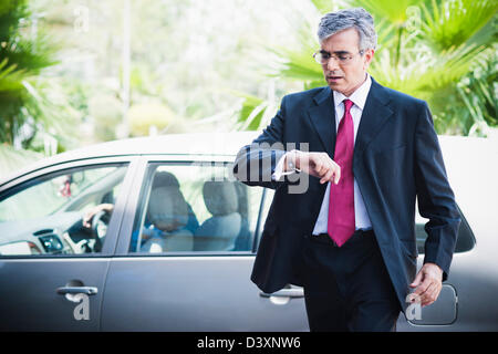 Imprenditore in piedi vicino a una macchina guardando il suo orologio da polso Foto Stock
