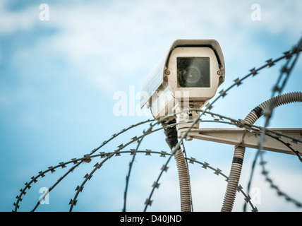 Vista ravvicinata di una telecamera di sicurezza appeso tra barbwire in prigione o altro oggetto custodito con cielo blu sullo sfondo. Metodi moderni Foto Stock