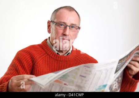 Uomo maturo in 50s che indossa un maglione, la lettura di un quotidiano broadsheet, guardando alla telecamera gravi. Foto Stock
