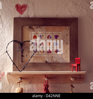 Close-up di filo spinato e di cuore in miniatura sedia rossa sul ripiano con peg rampa e telaio di legno intorno a cuori stampigliato sulla parete Foto Stock