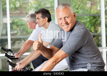 Felice l'uomo anziano in bici tenendo le dita fino in centro fitness Foto Stock