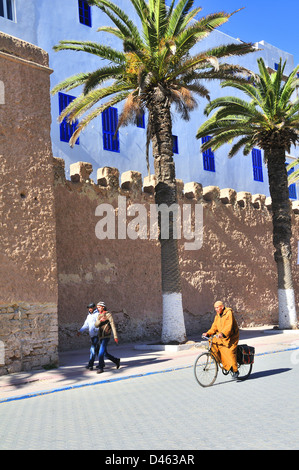 Uomo marocchino in camice giallo pedalando lungo la strada con due palme, mura fortificate e l'alloggio nella città costiera atlantica di Essaouira, Marocco Foto Stock