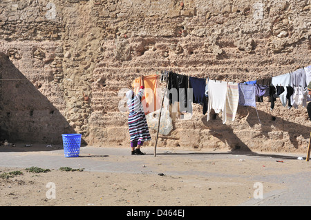 La donna a Essaouira, Marocco, appendere il suo esterno di lavaggio a secco sulla linea dietro alto muro di mattoni con il mare oltre Foto Stock