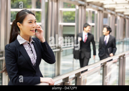 Imprenditrice cinese al di fuori ufficio su telefono cellulare con i colleghi in background Foto Stock