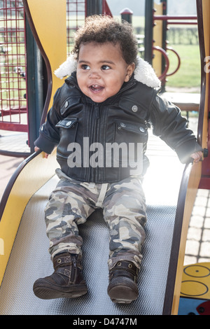 Razza mista toddler baby indossando giacca aviator giocando sulla parte superiore della diapositiva ridendo nella luce del sole Foto Stock