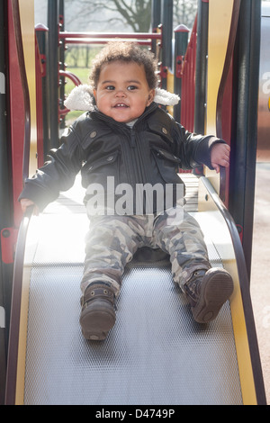 Razza mista toddler baby indossando giacca aviator giocando sulla parte superiore della diapositiva sorridente nella luce del sole Foto Stock