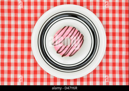 Una ciambella con glassa rosa e una spruzzata di cioccolato, su una piastra bianca che è collocato su un rosso-e-bianco tovaglia a scacchi, studio shot preso, vista dall'alto. Foto Stock