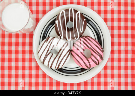 Decorate ciambelle glassate su una piastra bianca, con un bicchiere di latte, su un rosso-e-bianco, a scacchi tovaglia da picnic, studio shot presi dall'alto. Foto Stock