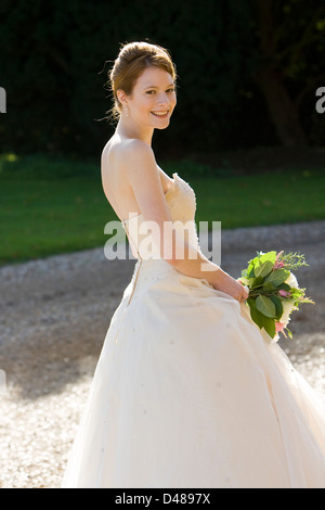 Sposa sorridente in piedi sul lato della telecamera nella luce del sole che indossa la crema senza spalline abito da sposa bouquet di contenimento Foto Stock