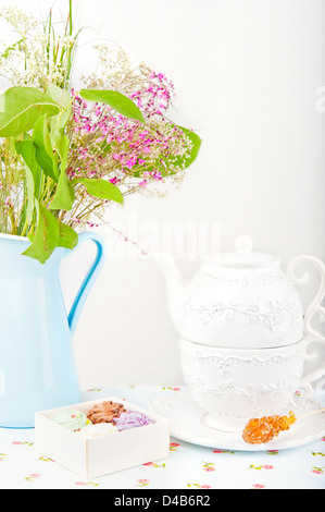 Tazza bianca e teiera, quattro caramelle colorate, zucchero e stick fiori in una brocca blu Foto Stock