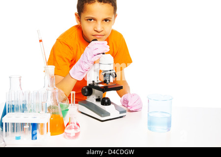 Felice black boy con capelli corti in laboratorio di chimica classe con microscopio e provette per la prova sul tavolo, isolato su bianco Foto Stock