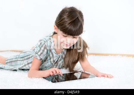 Bambina gioca con una moderna digitale compressa mentre giaceva su un tappeto bianco in una camera bianca Foto Stock