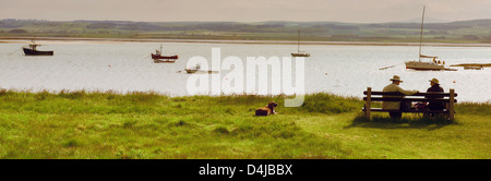 Vecchia coppia con cane rilassante su banco affacciato sulla Baia di Isola Santa, Northumberland Foto Stock